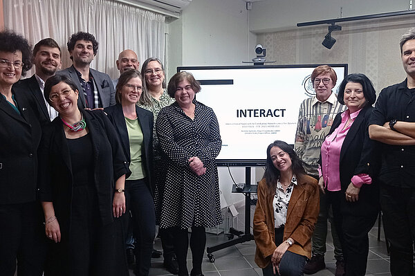 Gruppenbild INTERACT Das Foto zeigt 12 Frauen und Männer stehend vor einem Bildschirm mit der Aufschrift INTERACT